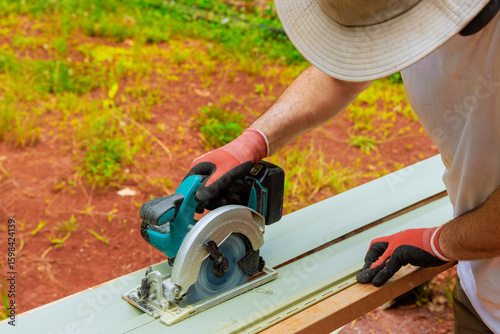 Carpenter is operating circular power saw on vinyl siding plank while working on construction project outside.