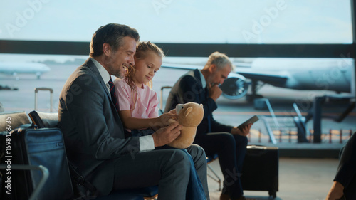 Obraz na plátne Business dad spends a heartfelt moment with his daughter at the airport, playing with her teddy bear while waiting for a flight — capturing work-life balance, fatherhood, and modern travel lifestyle