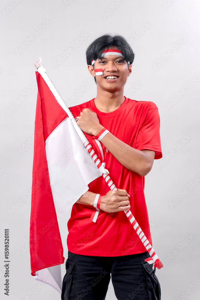 A smiling young Indonesian man proudly holds the Indonesian flag, displaying his patriotism with face paint and wristbands. He poses with excitement, symbolizing national pride. A smiling young Indonesian man proudly holds the Indonesian flag, displaying his patriotism with face paint and wristbands. He poses with excitement, symbolizing national pride.