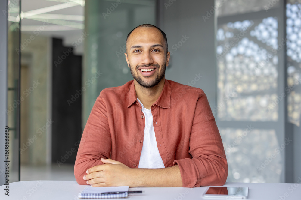Naklejka premium A smiling man sits at a table, appearing confident and relaxed. The setting suggests a modern office or workspace.