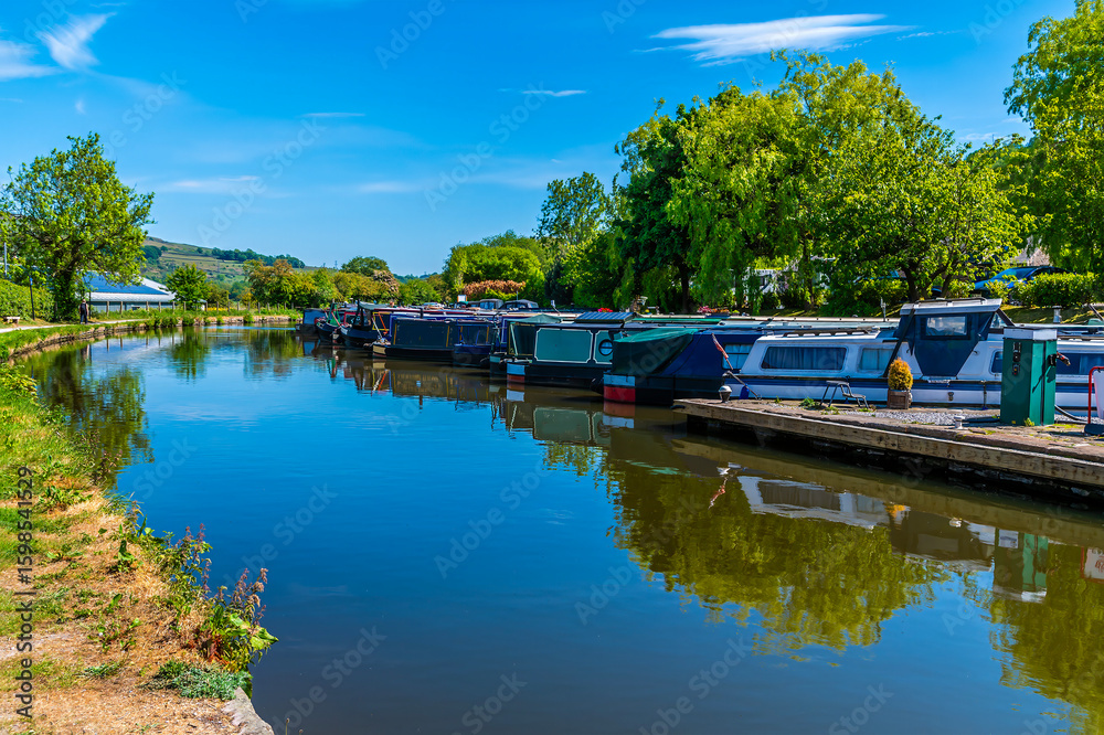 Obraz premium A view down the Peak Forest Canal past moored canal barges at New Mills, High Peak in the UK in summertime