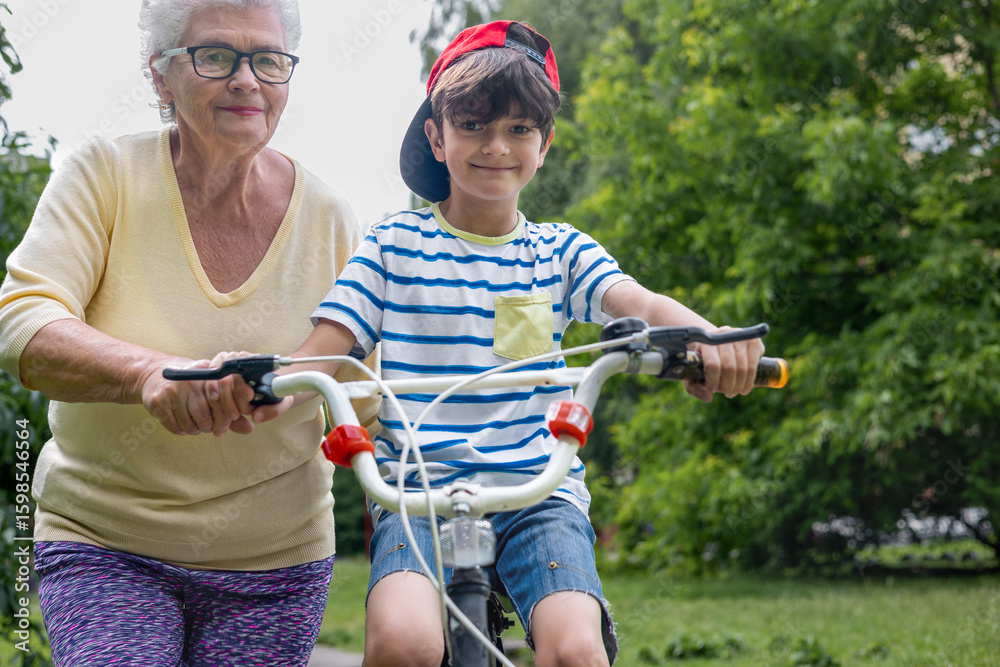 Obraz premium Grandmother Guides Grandson Learning to Ride Bicycle with Training Wheels. Family Bonding, Child Development, Active Summer Outdoor Activity. Copyspace