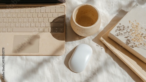 A minimalist workspace featuring a laptop, a white mouse, a wooden pot, and a notebook on a light fabric surface. Natural light creates a warm atmosphere.
