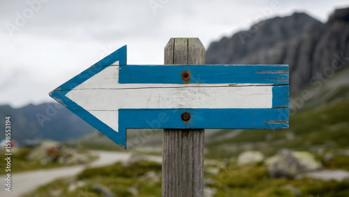 Weathered wooden signpost with a blue and white arrow pointing left against a blurred mountain landscape, symbolizing direction and choice