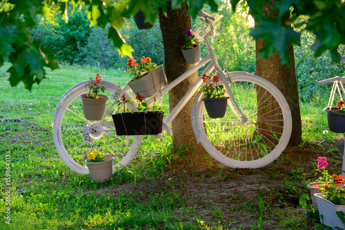 White vintage bicycle decorated with colorful flower pots used as a creative garden decor, standing under a tree in summer park, eco-friendly outdoor decoration with bright floral arrangement