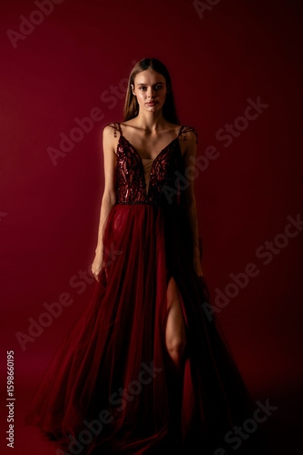 Elegant woman in deep red gown standing against maroon backdrop showcasing fashion and grace during a studio photoshoot