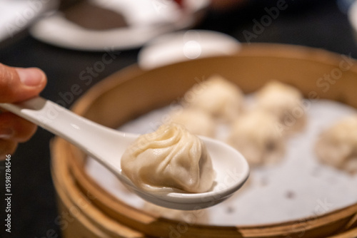 Close-up of a single xiao long bao resting on a spoon, lifted from a round bamboo steamer in the background