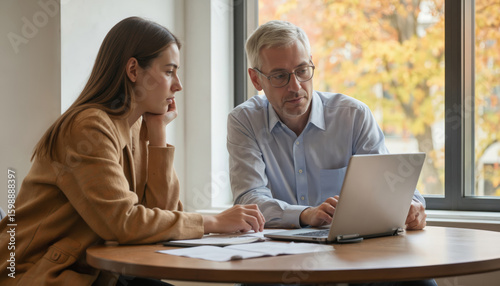University professor consults with graduate student discussing project materials on laptop. Woman student listens attentively. Man teacher offers guidance for academic growth, career development,
