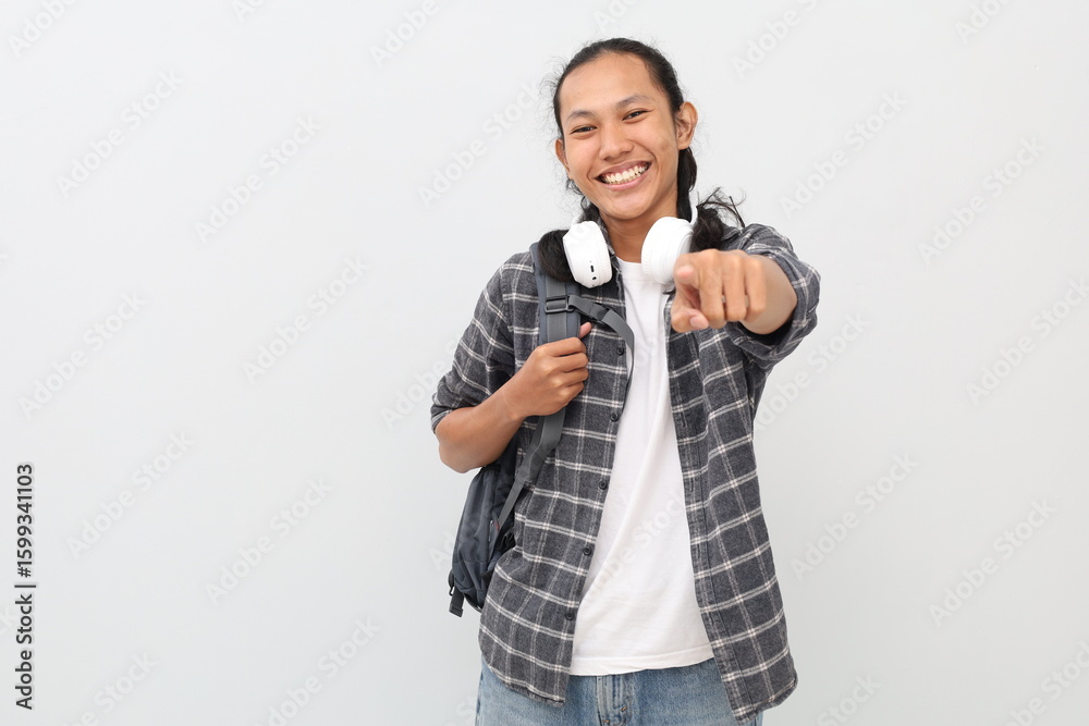 Young Asian male student wearing casual clothes and backpack, headphones around neck, pointing forward, standing smiling looking at camera. Young Asian male student wearing casual clothes and backpack, headphones around neck, pointing forward, standing smiling looking at camera.