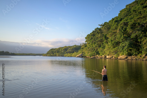 Cena tranquila de um menino pescando sozinho na margem de um rio, em meio à natureza exuberante da mata atlântica. Simplicidade, conexão com a natureza e infância livre.
