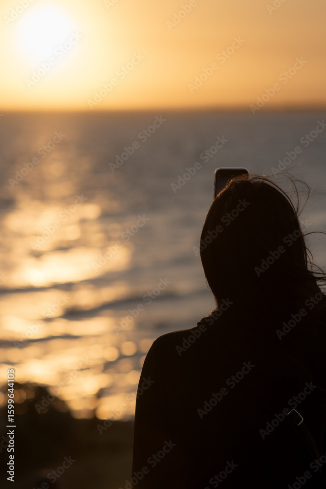 Fototapeta premium Woman taking photo of golden sunset over ocean with phone