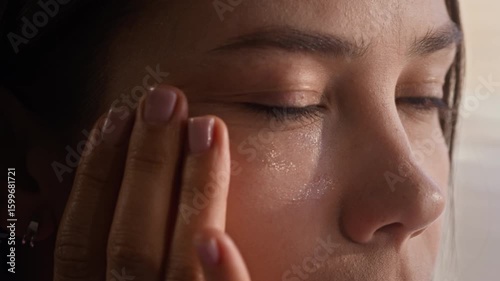 Close-up of young beautiful woman applying hydrating gel on perfect skin of her face and looking at herself in mirror while tapping cosmetic product with fingertips