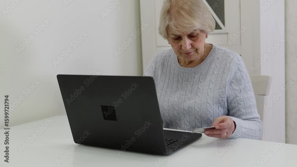 Concentrated senior woman managing personal finances at home, securely processing online transaction using laptop and credit card while sitting at white table