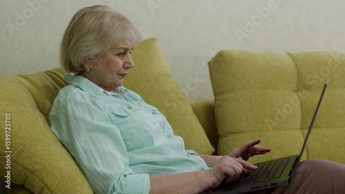 Focused elderly woman sitting comfortably on a sofa and typing on a laptop, potentially working remotely or engaging in online communication