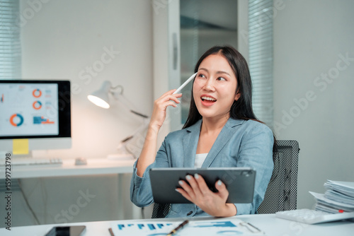 Young businesswoman sitting at a desk in a modern office, having a bright idea while using a tablet and holding a pen thoughtfully near her head, exuding confidence and creativity