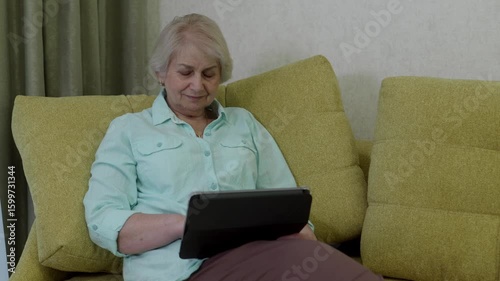 Cheerful retired woman relaxing on comfortable sofa, exploring digital tablet, connecting with friends and enjoying modern technology in cozy home environment
