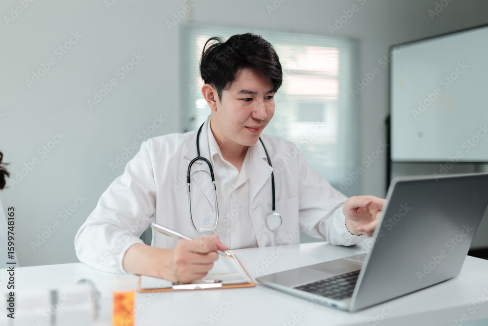 Young male physician wearing a lab coat and stethoscope, sitting at a desk in a bright medical office, using a laptop and taking notes on a clipboard while working diligently Young male physician wearing a lab coat and stethoscope, sitting at a desk in a bright medical office, using a laptop and taking notes on a clipboard while working diligently