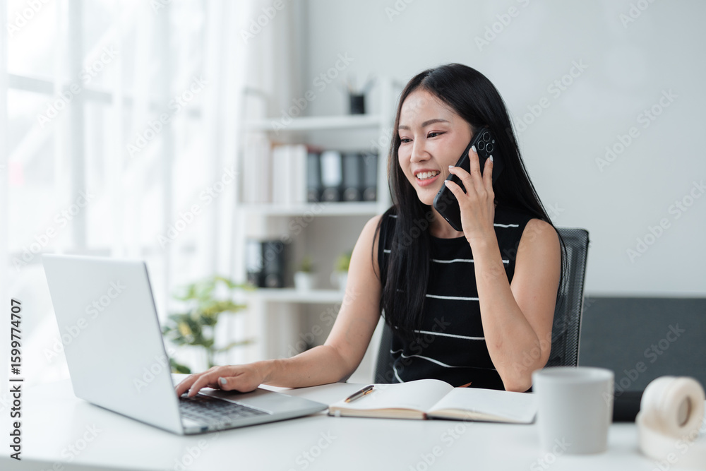 Young Asian businesswoman is multitasking in a bright office, talking on her smartphone while working on her laptop, showcasing efficiency and connectivity in the modern workplace Young Asian businesswoman is multitasking in a bright office, talking on her smartphone while working on her laptop, showcasing efficiency and connectivity in the modern workplace