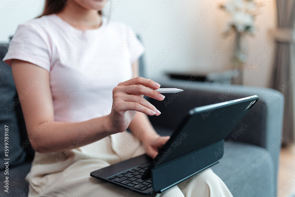 Fototapeta premium Young woman sitting comfortably on a sofa, using a tablet with a keyboard and stylus while working or studying from home in a cozy, well lit living room