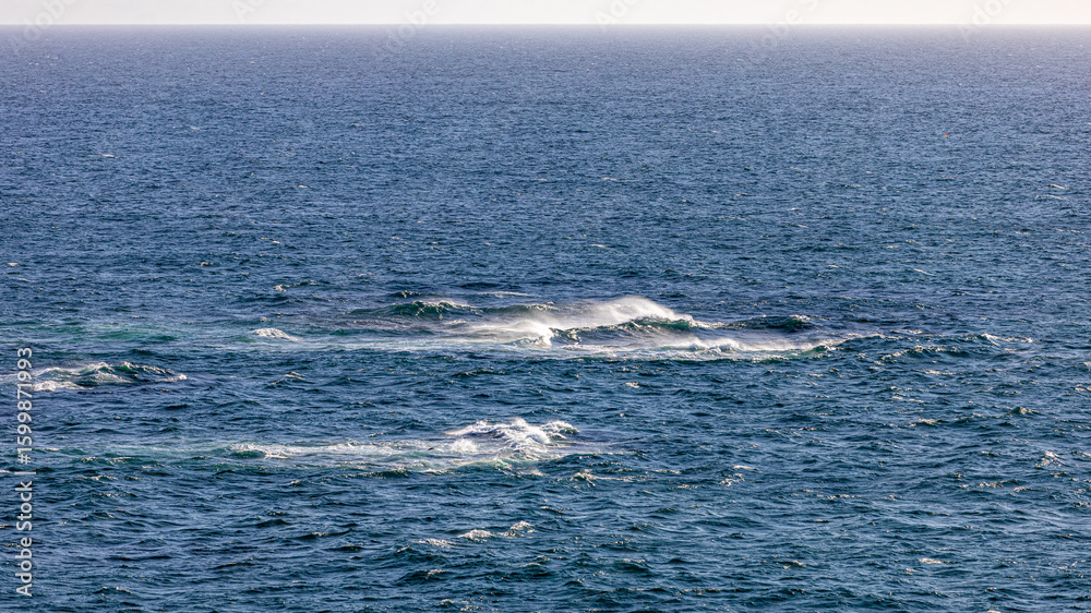 Fototapeta premium A dynamic ocean scene with waves crashing over submerged rocks, sending sea spray into the air under clear skies. Perfect for travel, nature, and seascape themes.