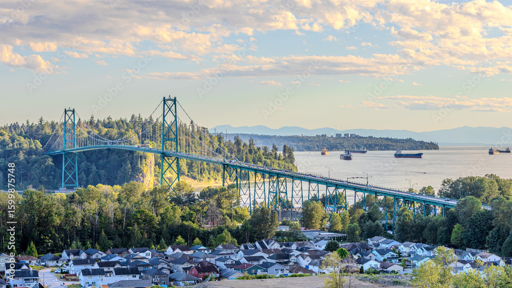 Fototapeta premium Panoramic view of the iconic Lions Gate Bridge spanning the inlet, with cargo ships, forested hills, and a residential neighborhood below at sunset.