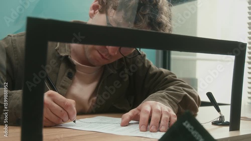 Shot from behind service window glass of male client signing document to withdraw money then receiving cash from bank teller at counter