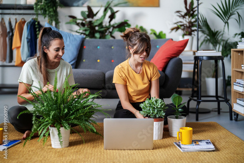 Two women are sitting on the floor in a cozy living room, surrounded by various houseplants. One woman tends to a large fern while the other looks at a laptop with smaller plants