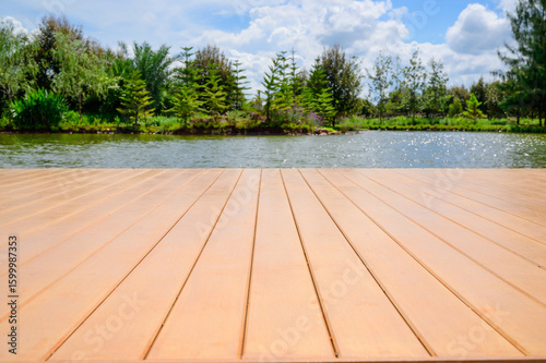 A serene outdoor scene is captured from the perspective of a light-colored wooden deck or pier, which occupies the lower half of the frame and leads towards a calm body of water.