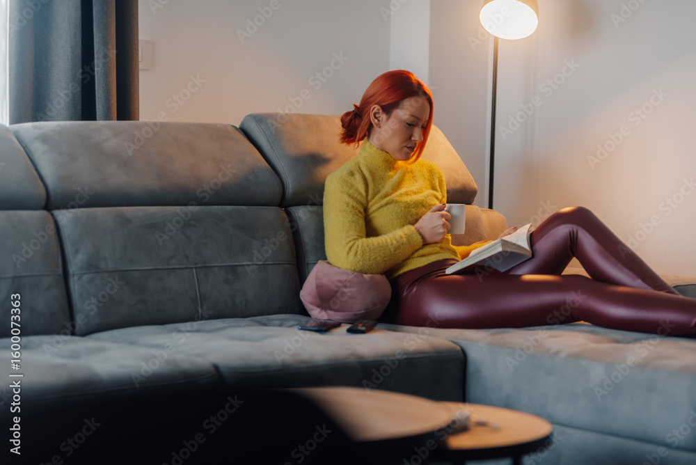 Obraz premium Woman relaxing on sofa reading a book and drinking tea