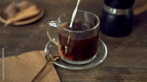 A stream of milk is being poured into a glass mug filled with rich coffee, creating beautiful swirling patterns. 