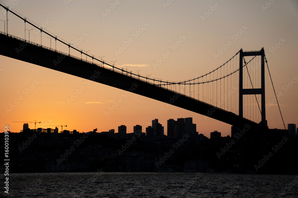 Obraz premium Suspension bridge silhouette at sunset with city skyline.