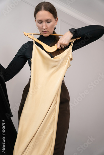 Young woman holding two dresses, one in luxurious black velvet and the other in soft beige silk, thoughtfully comparing options while deciding on the perfect outfit for the occasion. Vertical photo