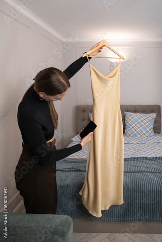 Fashion designer holding a beige slip dress on a hanger and taking photos of it with her smartphone, standing in a bedroom with a bed in the background, showcasing her latest creation