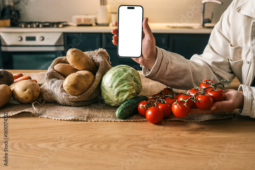 Person holding a smartphone with blank white screen over a rustic kitchen table filled with fresh vegetables: potatoes, tomatoes, cabbage, cucumber. Mockup concept with natural lighting