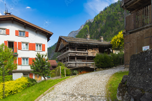 A charming cobblestone street in Sottoguda, a traditional Alpine village, flanked by historic wooden and stucco houses with lush green mountains in the background.