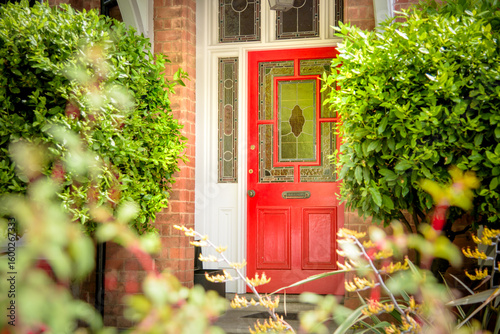 Welcoming vibrant red front door in English red brick residential house. Inviting home entrance. Charming victorian doorway with beautiful stained glass sidelight and transom windows and front garden.