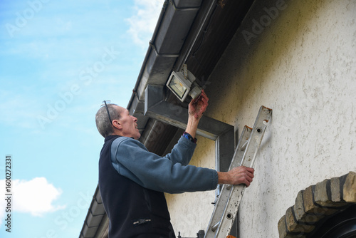 Electrician adjusts and controls motion sensor LED floodlight while standing on ladder,light fixture on facade of house improve outdoor lighting when needed, saving energy during energy crisis,