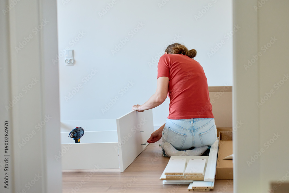 Woman sits on wooden floor and assembling furniture in new home, fastens parts of white bed frame. DIY home renovation and home organization after moving concept Woman sits on wooden floor and assembling furniture in new home, fastens parts of white bed frame. DIY home renovation and home organization after moving concept