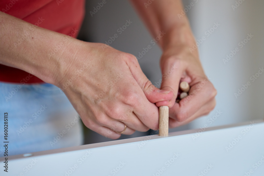 Female hands insert wooden dowel into white furniture board during assembly. Woman connects furniture parts using wooden pegs