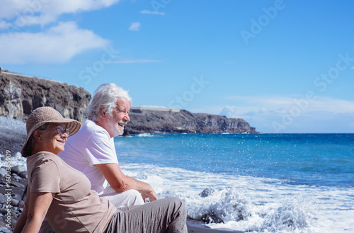 Relaxed senior couple in peaceful vacation sitting on the seashore while waves splashing on the beach enjoying retirement lifestyle and freedom, horizon over sea