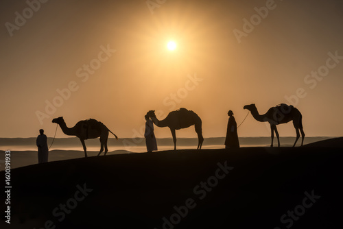 Wahiba Sands, Oman - 12 May 2022: View of a silhouetted camel caravan led by robed figures cresting a dune under the soft glow of the desert sun.
