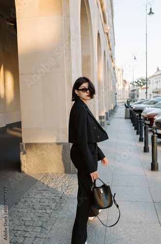 Fotografie Confident woman in black pantsuit walking through city street during golden hour