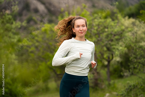 Portrait of young smiling woman running jogging in mountains in daylight, doing sports exercises in nature, wearing activewear leggings and long sleeve, outdoor activities in summertime