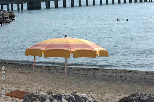 Ombrellone giallo-arancio e lettino vuoto su una spiaggia di ciottoli del Mediterraneo con sfondo del molo.