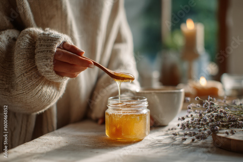 woman in cozy sweater holding spoonful of golden honey over jar on rustic table with herbs and candles, warm morning light, concept of homestead core, natural wellness, slow living