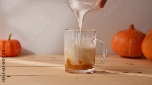Steamed milk flows into glass mug with pumpkin syrup and ice, creating a vibrant homemade pumpkin latte with warm autumn atmosphere.