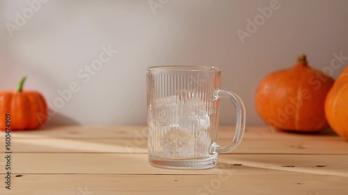 Iced pumpkin latte preparation in glass mug with ice and milk, autumn pumpkins on wooden table, cozy seasonal atmosphere.