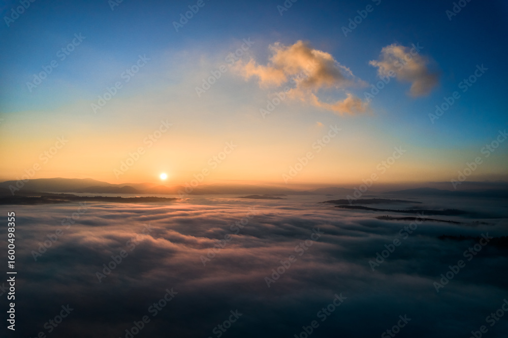 Fototapeta premium Aerial view of sun rises above dense clouds, casting warm, golden glow across horizon. Scene captures tranquility of new day, as the first rays of sunlight break through, creating serene atmosphere.