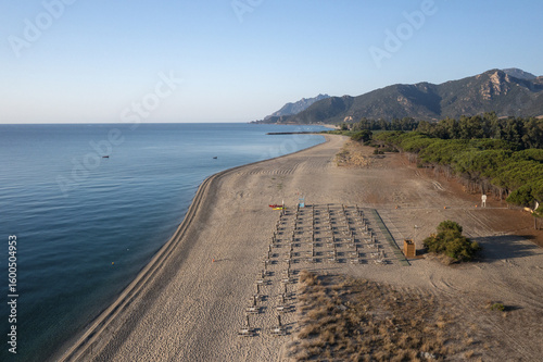 Aerial view of the golden beach meeting the turquoise sea, with rows of sunbeds awaiting visitors under the clear sky, Nuoro, Sardinia, Italy.