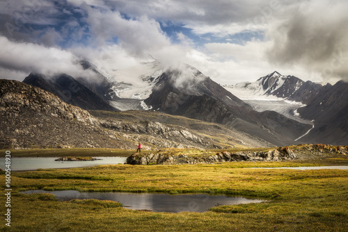 View of vibrant green meadows meet rugged mountains capped with snow under a dramatic sky, inviting exploration and wonder, Kyrgyzstan.